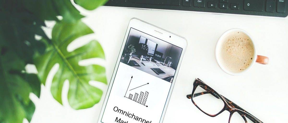 Mobile phone with Omnichannel Marketing on screen set on desk next to keyboard, eyeglasses, cup of coffee, and green plant